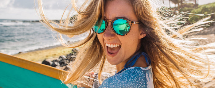 A woman smiling on a beach wearing a pair of Sunski mirrored sunglasses