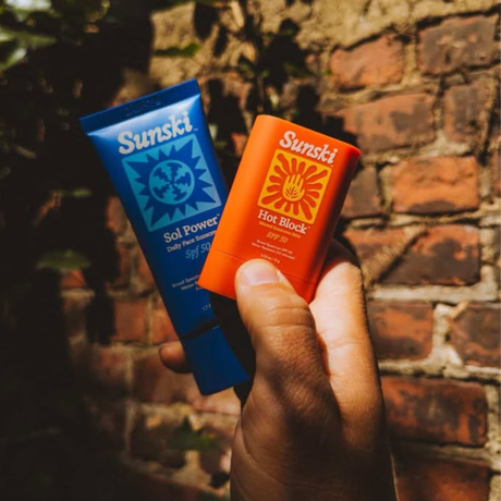 Hand holding two Sunski sunscreen products against a brick wall.