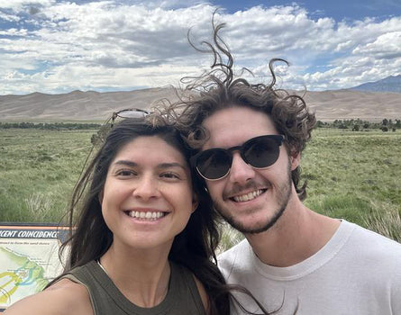 Two people posing for a selfie with a scenic background of mountains and open fields.