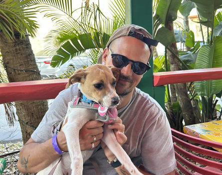 Man holding a small dog outdoors with greenery in the background