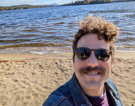 Man with sunglasses and a mustache standing on a beach with water and sky in the background