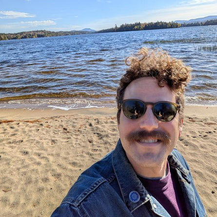 Man with sunglasses and a mustache standing on a beach with water and sky in the background