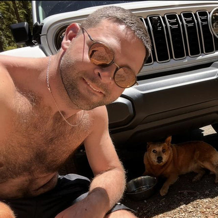 Man with sunglasses and a dog sitting next to a Jeep