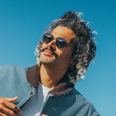 A man with curly gray and white hair wearing round sunglasses and a striped jacket with a brown sherpa collar, photographed against a clear blue sky.