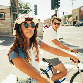 A woman in a cream and brown knit hat and sunglasses rides a bicycle in a sunny street scene, with a man on a white bicycle behind her.
