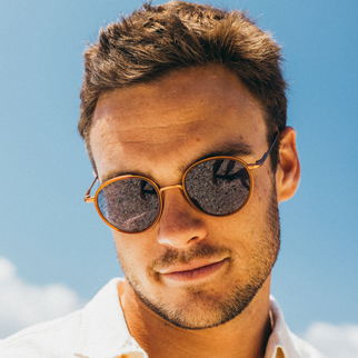 A close-up portrait of a man wearing round gold-framed sunglasses with dark mirrored lenses, set against a bright blue sky background.