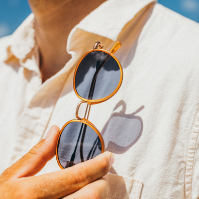 A close-up of a person wearing a light beige button-up shirt, holding a pair of round gold-framed sunglasses with dark lenses in their hand.