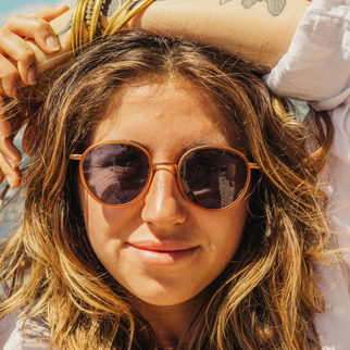 A close-up lifestyle shot of a woman with wavy light brown hair wearing round copper-toned sunglasses with dark lenses, holding a bottle near her head.