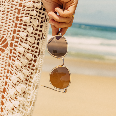 A person at the beach holds a pair of round gold-framed sunglasses with brown tinted lenses, wearing a cream crochet cover-up dress.