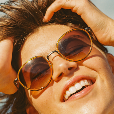 A close-up lifestyle shot of a smiling woman outdoors wearing round tortoiseshell sunglasses with dark tinted lenses.