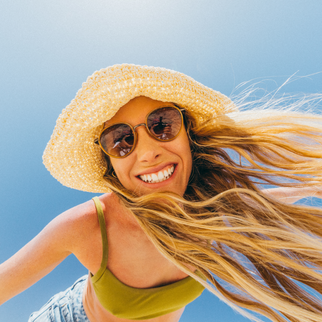 A smiling woman outdoors against a clear blue sky, wearing a large woven straw hat, round sunglasses, a green top, and denim shorts, with her hair blowing in the wind.