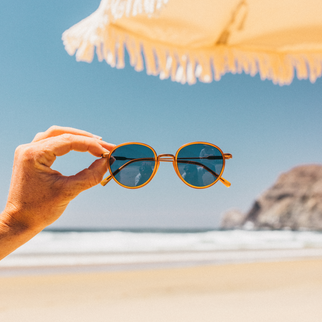 A hand holds a pair of round amber-framed sunglasses with dark blue lenses against a sunny beach background with a blurred umbrella and ocean.