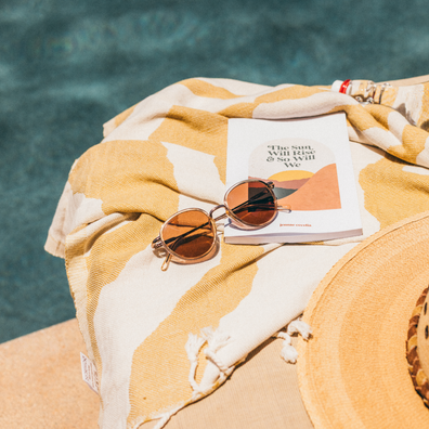 A styled flat-lay of a yellow and cream striped woven towel with tassels, featuring a book, round sunglasses, and a straw hat, set next to a swimming pool.