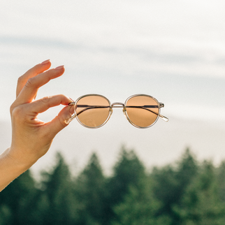 A hand holding a pair of round sunglasses with gold metal frames and warm brown-tinted lenses against a soft-focus outdoor background of a cloudy sky and green trees.