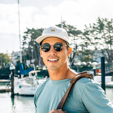 A smiling man wearing a white baseball cap with a small gold logo, dark sunglasses, and a light blue t-shirt stands at a sunny marina with boats and trees in the background.