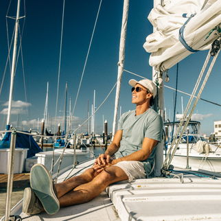 A man wearing a light green t-shirt, white shorts, a white cap, and sunglasses sits relaxed on the deck of a white sailboat at a sunny marina under a deep blue sky.