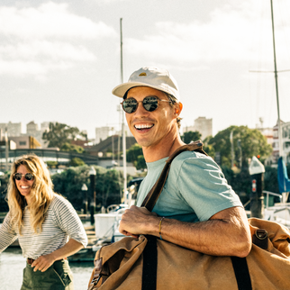 A smiling man in a light blue t-shirt, beige cap, and sunglasses carries a large tan duffel bag on a harbor with a woman in the background.