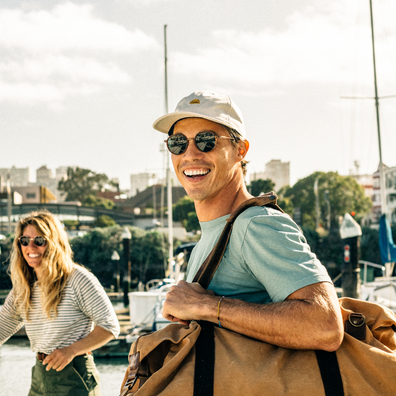 A smiling man in a light blue t-shirt, beige cap, and sunglasses carries a large tan duffel bag on a harbor with a woman in the background.
