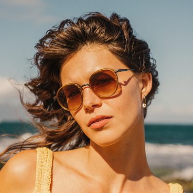 A woman outdoors by the beach wearing round amber-tinted sunglasses with a gold frame and a small gold hoop earring.