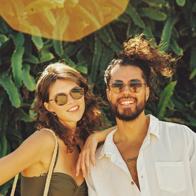 A smiling couple posing outdoors in front of green cactus foliage. The woman on the left wears round sunglasses and a dark olive strapless top, while the man on the right wears aviator-style sunglasses and an open white button-down shirt.