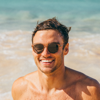 A smiling man on a beach wearing round sunglasses with a gold-tone frame and dark lenses, with turquoise ocean water in the background.