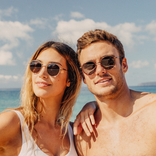 A man and a woman wearing round dark sunglasses with gold-toned frames, posing together on a sunny beach with the blue ocean and sky in the background.