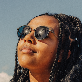 A close-up lifestyle shot of a woman with braided hair wearing round metal-framed sunglasses with dark lenses reflecting a blue sky and palm trees.