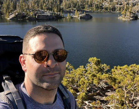 Man with sunglasses and backpack standing in front of a lake and forest