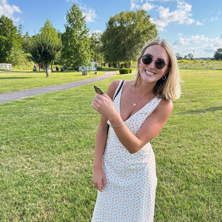 Woman in a white dress holding a phone in a park with green grass and trees.