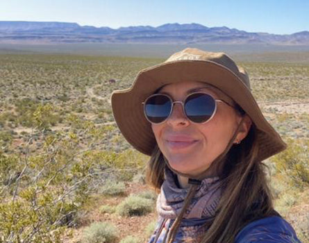 Person wearing a wide-brimmed hat and sunglasses in a desert landscape with mountains in the background.