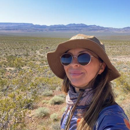 Person wearing a wide-brimmed hat and sunglasses in a desert landscape with mountains in the background.