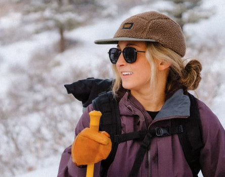 Person hiking in a snowy landscape wearing a brown cap, sunglasses, and purple jacket.