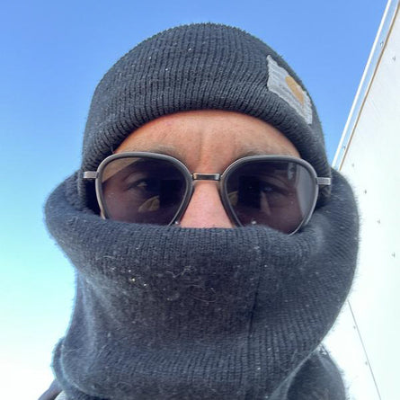 Person wearing a gray knit hat and balaclava with sunglasses, against a blue sky.