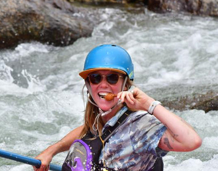 Person wearing a blue helmet and sunglasses, holding a paddle in turbulent water with rocks.