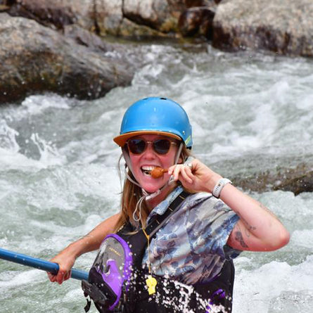 Person wearing a blue helmet and sunglasses, holding a paddle in turbulent water with rocks.