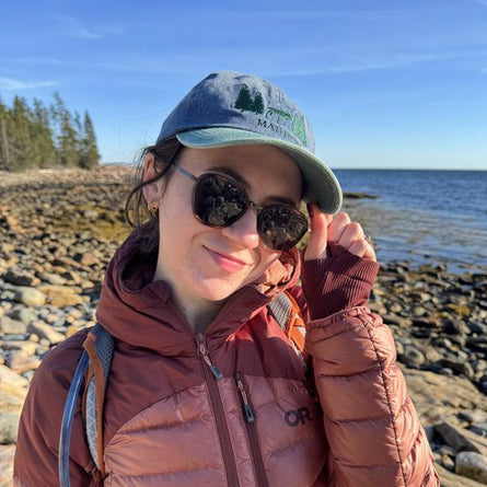 Person wearing sunglasses and a cap by a rocky shoreline with water and sky in the background