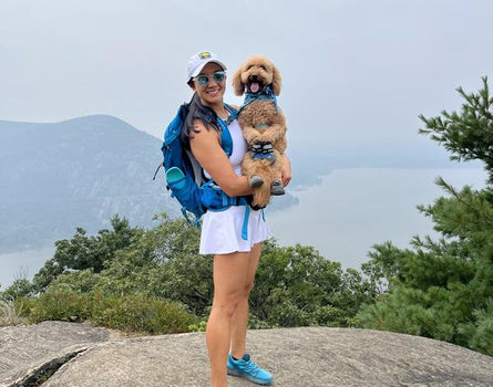 Woman with a dog on her arm standing on a rocky outcrop with mountains in the background