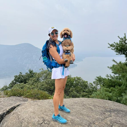 Woman with a dog on her arm standing on a rocky outcrop with mountains in the background