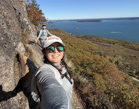 Person taking a selfie on a rocky cliff overlooking a lake with another person in the background.