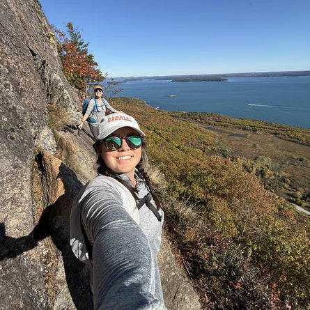 Person taking a selfie on a rocky cliff overlooking a lake with another person in the background.