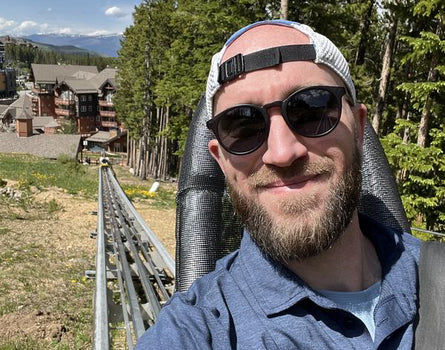 Man with sunglasses and a beanie standing outdoors with trees and buildings in the background