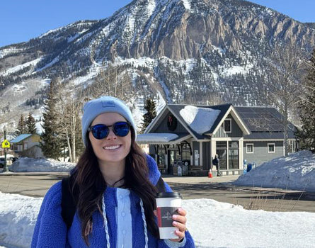 Person in blue coat and sunglasses holding a coffee cup with a mountain and building in the background