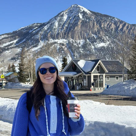 Person in blue coat and sunglasses holding a coffee cup with a mountain and building in the background