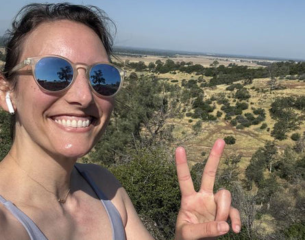 Woman wearing sunglasses and a gray tank top, making a peace sign with her hand, with a scenic landscape in the background.
