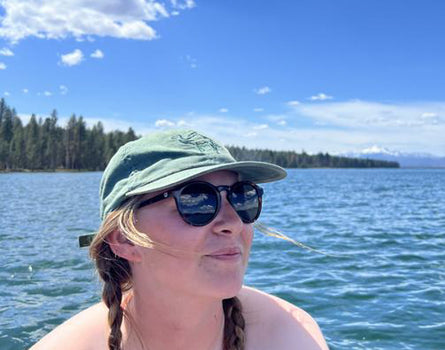 Person wearing sunglasses and a cap by a lake with trees in the background