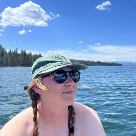 Person wearing sunglasses and a cap by a lake with trees in the background
