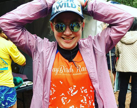 Person wearing a purple jacket and orange shirt with reflective sunglasses and a cap, standing outdoors.