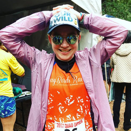 Person wearing a purple jacket and orange shirt with reflective sunglasses and a cap, standing outdoors.