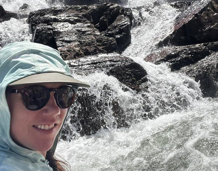 Person wearing a light blue hooded jacket and sunglasses in front of a flowing river with rocks.