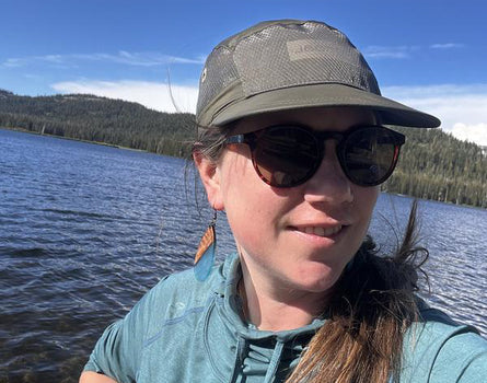 Person wearing sunglasses and a cap by a lake with mountains in the background